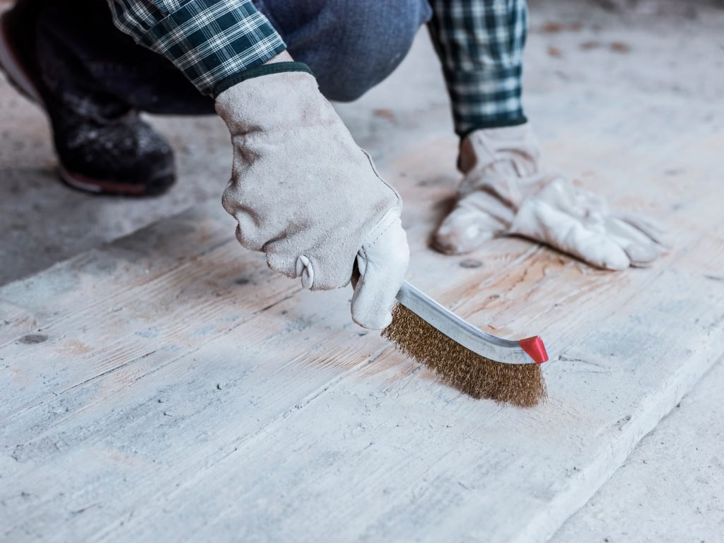 A gloved bricklayer uses a wire brush to remove cement residue from a wooden floor.